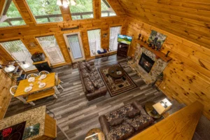Overhead view of cabin living room with pine walls, fireplace, leather seating, dining table, and forest views