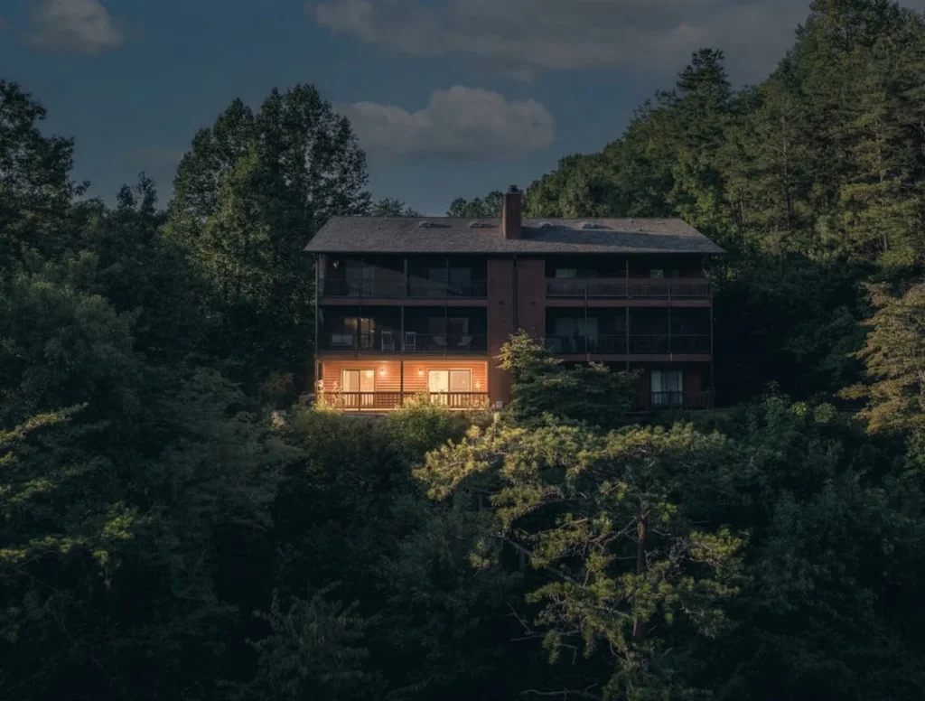 Modern multi-story cabin in Gatlinburg Tennessee forest with glowing windows, cedar siding, and stone chimney at dusk